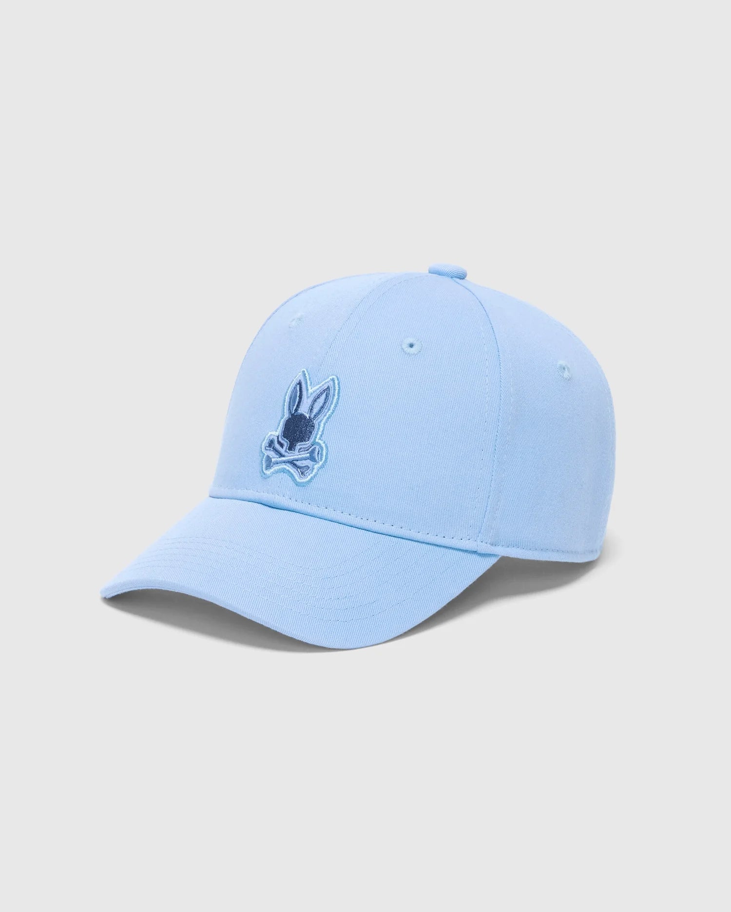 The Psycho Bunny Men's New Castle Embroidered Graphic Cap (B6A547F200) is a light blue cotton twill baseball cap. It features a small embroidered cartoon rabbit face with crossed bones, showcasing quiet luxury with a curved brim on a plain white background.
