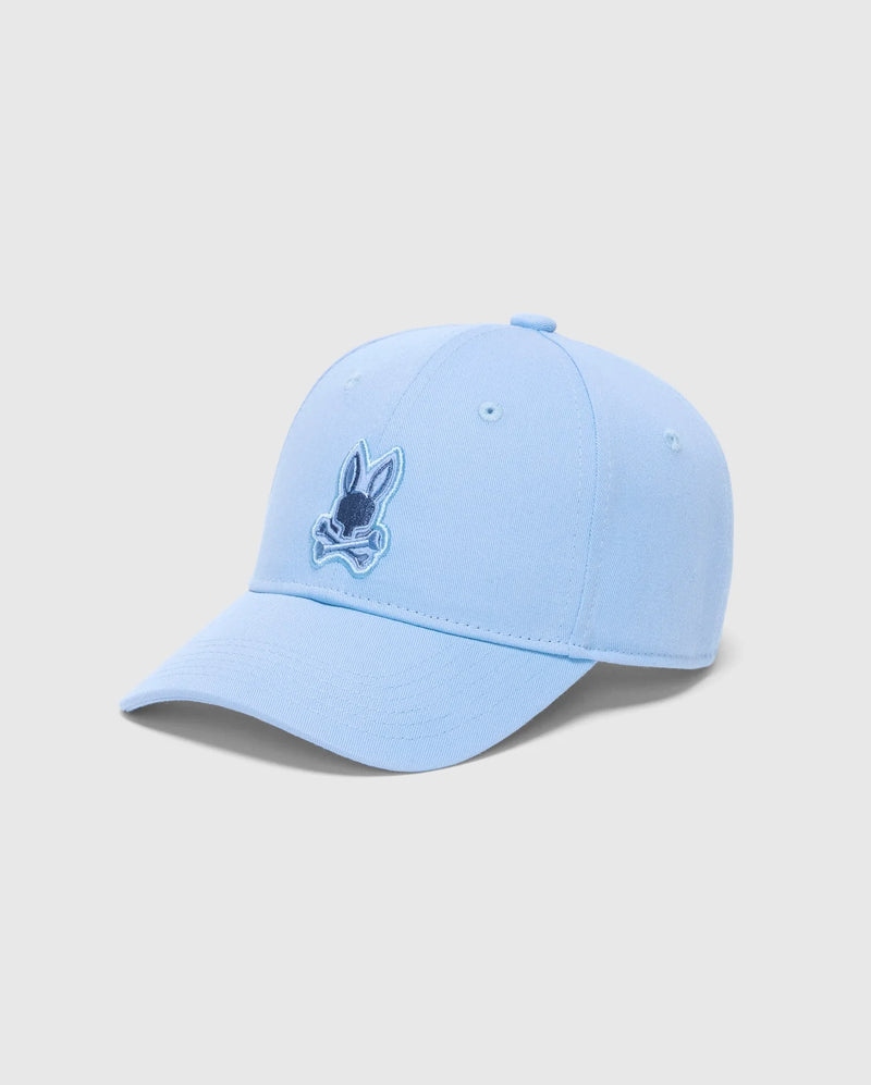 The Psycho Bunny Men's New Castle Embroidered Graphic Cap (B6A547F200) is a light blue cotton twill baseball cap. It features a small embroidered cartoon rabbit face with crossed bones, showcasing quiet luxury with a curved brim on a plain white background.
