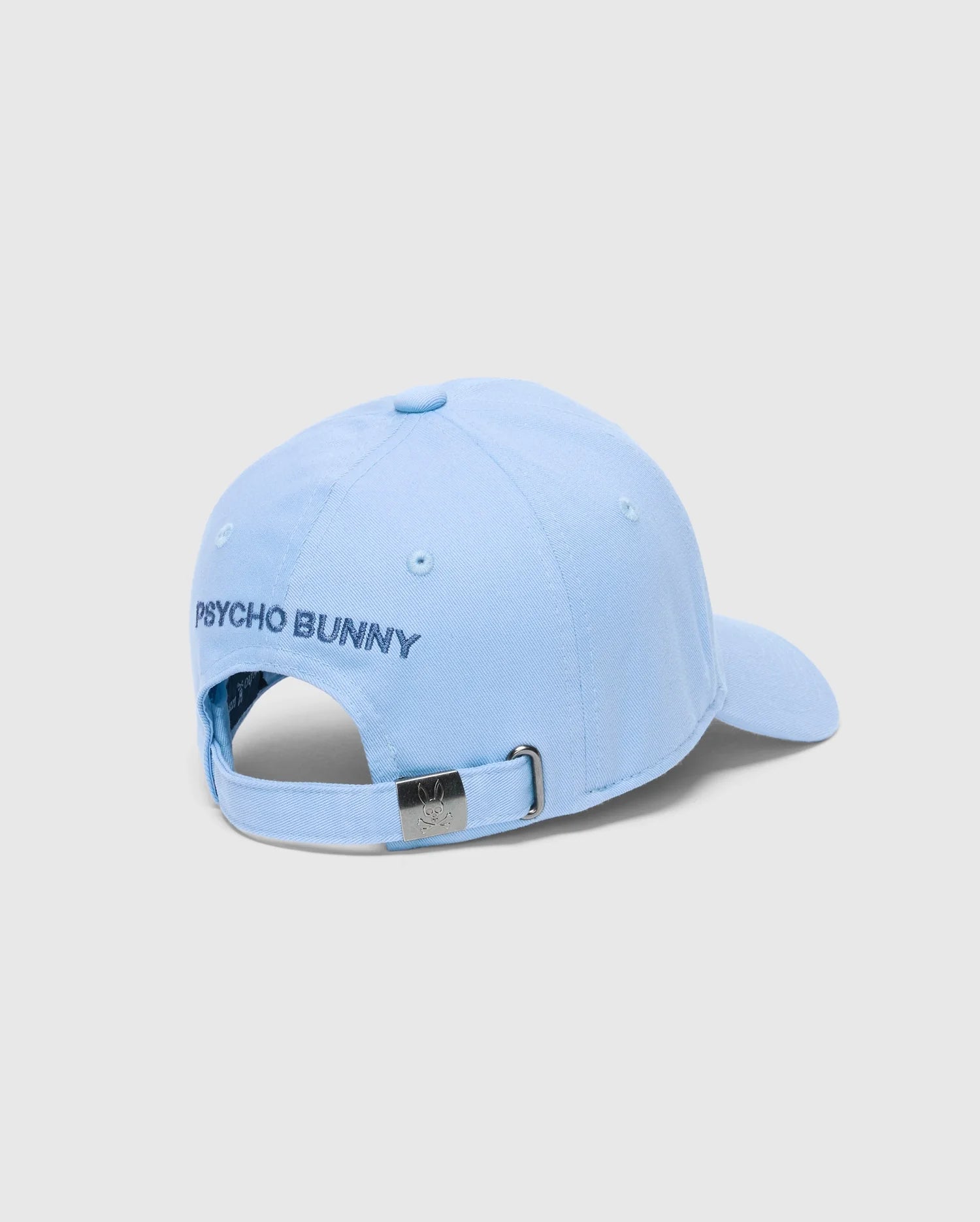 The MENS NEW CASTLE EMBROIDERED GRAPHIC BASEBALL CAP - B6A547F200 from Psycho Bunny is a light blue cotton twill cap with "Psycho Bunny" in dark blue on the back, an adjustable strap with a metal buckle, and a small metal logo above it.