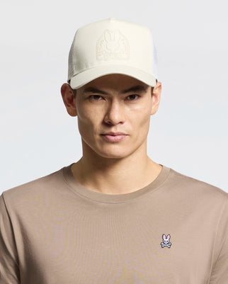 A person wearing a Psycho Bunny MENS MELBOURNE GRAPHIC TRUCKER HAT - B6A548F200 with a subtle logo and embroidered icon, paired with a taupe T-shirt featuring a small pink bunny emblem, stands against a plain light gray backdrop.
