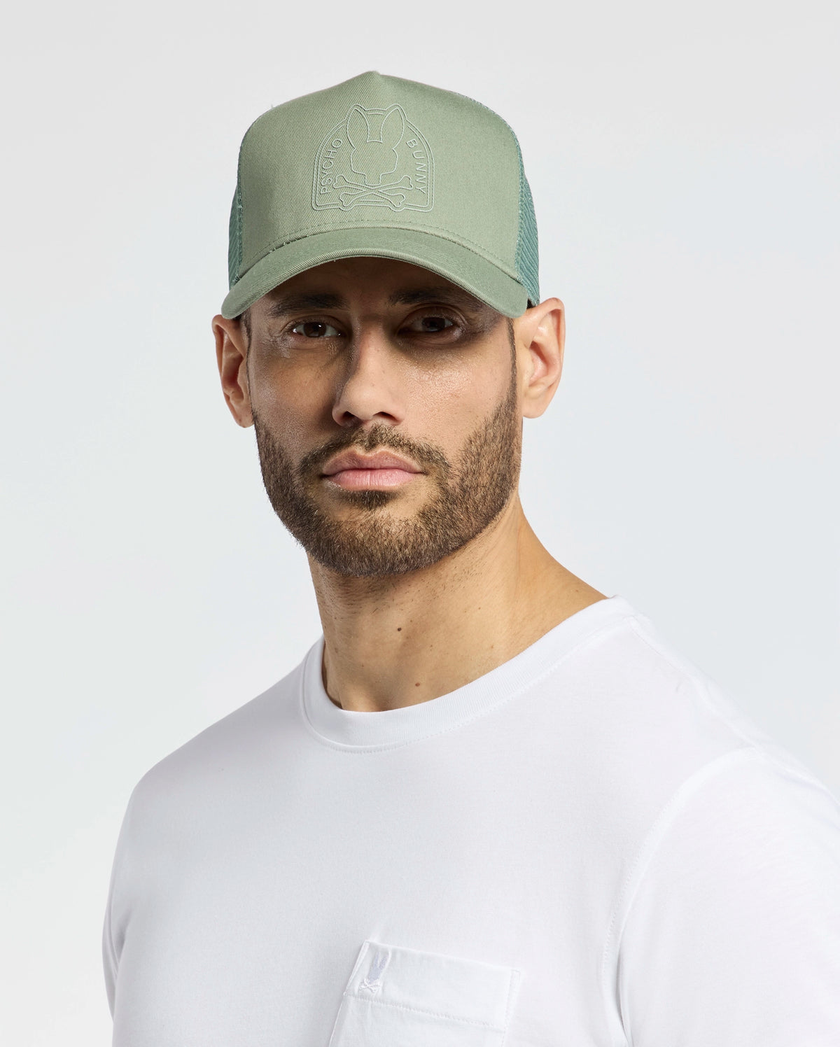 A bearded individual wears a breathable light green Psycho Bunny Melbourne Graphic Trucker Hat with a subtle emblem and snap-back strap, paired with a white pocket t-shirt on a plain background.