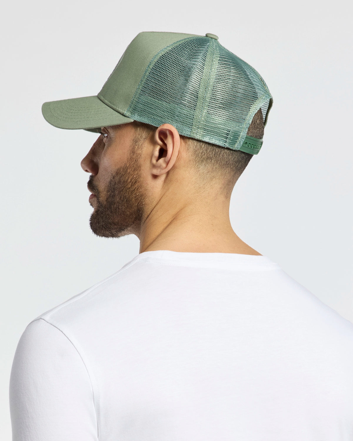 A person wearing Psycho Bunny's MENS MELBOURNE GRAPHIC TRUCKER HAT - B6A548F200 with a green mesh-back and breathable design, paired with a white shirt, is facing away towards the left against a plain, light-colored background.