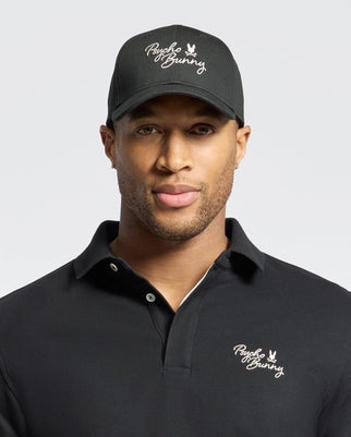 Against a plain backdrop, a man in a black cotton twill Psycho Bunny MENS DESERT BASEBALL CAP with an adjustable strap and a polo shirt displaying the brand's distinctive bunny logo maintains a neutral expression.