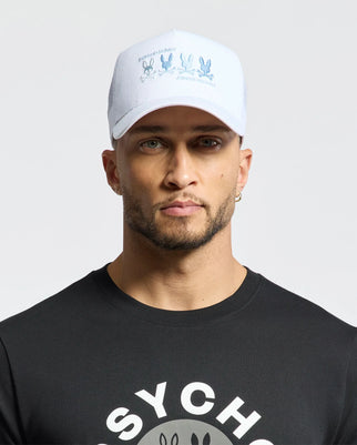 A person sports a Psycho Bunny Oceania Embroidered Graphic Trucker Hat and a black shirt with "Psycho Bunny" text, showcasing their streetwear style with a neat beard against a plain white background.