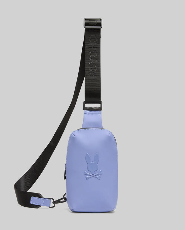 The Psycho Bunny MENS RUBBERIZED CROSSBODY (B6A635F200) is a small, rectangular light purple polyester bag with a black adjustable strap, embossed skull and bunny logo on the front, and "PSYCHO" text on the strap. Plain gray background.