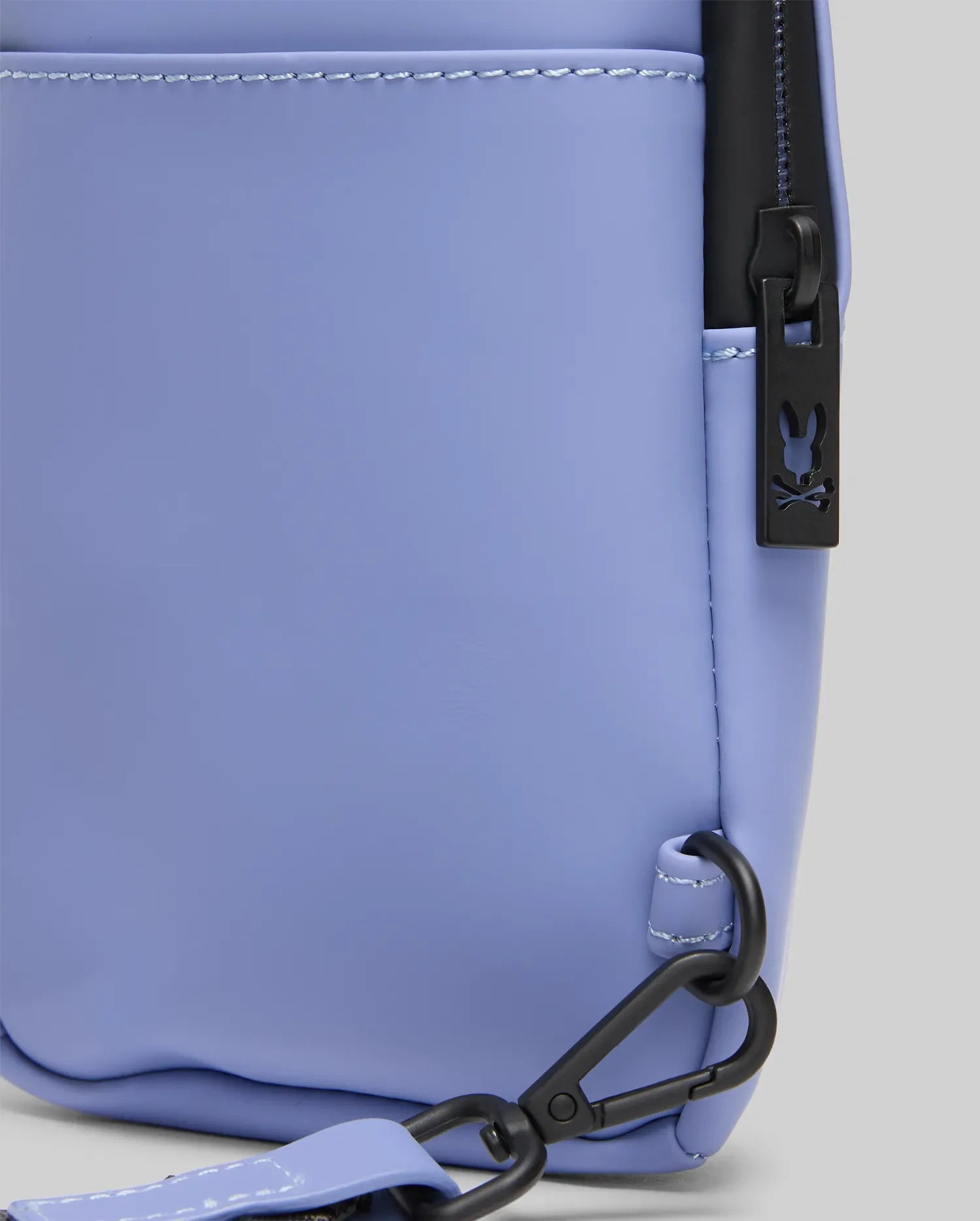 Close-up of the MENS RUBBERIZED CROSSBODY - B6A635F200 by Psycho Bunny in light purple polyester, featuring black zipper hardware, a bunny logo pull, and a matching adjustable strap with a black clasp attached to the bottom corner.