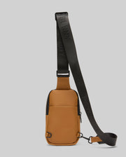 The Psycho Bunny MENS RUBBERIZED CROSSBODY - B6A635F200 is a small tan rectangular bag made from durable polyester, featuring a front pocket, black zipper, and wide embossed adjustable strap, shown upright against a light gray background.