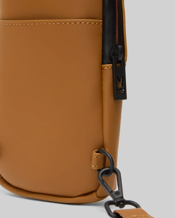 Close-up of the Psycho Bunny MENS RUBBERIZED CROSSBODY - B6A635F200 in tan polyester with a black zipper, small bunny logo, black hardware, and an adjustable detachable strap against a light gray background.