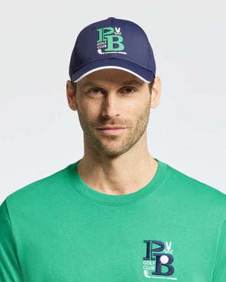 A man sports the MENS DENIS SPORT CAP by Psycho Bunny in navy, with "P B Golf Club" in green and white and an adjustable clasp. His green t-shirt, featuring the same logo on a lightweight cotton blend, stands out against a plain white background.