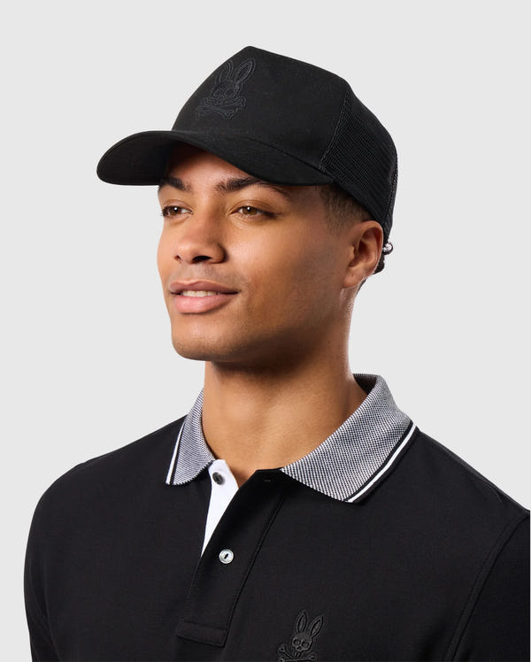 A man in a Psycho Bunny MENS MALTON TRUCKER CAP - B6A797D200, with mesh ventilation and bunny logo, wears a black polo shirt with white trim and smiles while looking to the side against a plain gray background.