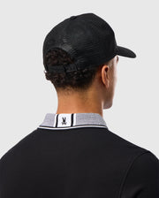 A person seen from the rear wears a Psycho Bunny MENS MALTON TRUCKER CAP - B6A797D200, offering mesh ventilation and an adjustable strap, with a black cotton polo shirt featuring a logo on the white and black designed collar. Light gray background.
