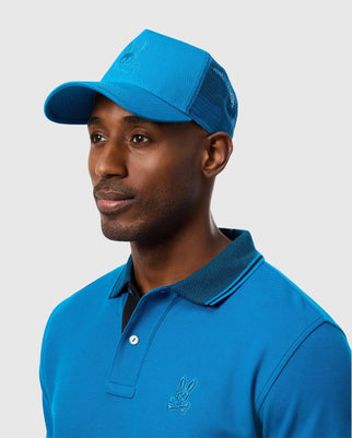 A man in a Psycho Bunny MENS MALTON TRUCKER CAP - B6A797D200 and a blue polo shirt stands against a light gray background, facing slightly to the side.