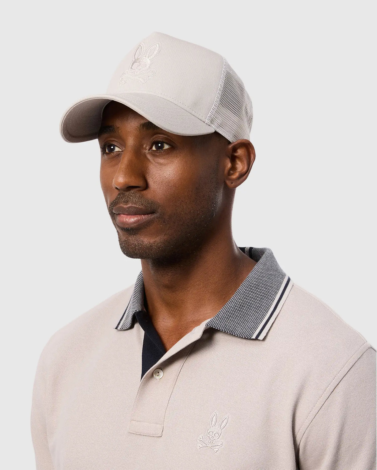 A person wearing a beige polo shirt and the Psycho Bunny MENS MALTON TRUCKER CAP - B6A797D200 with a bunny logo. The cap has mesh ventilation for comfort. The background is a plain light gray.
