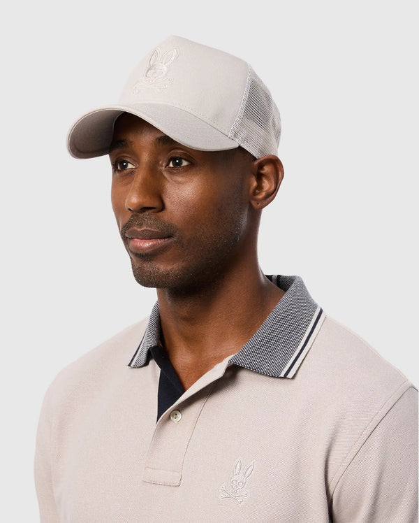 A person wearing a beige polo shirt and the Psycho Bunny MENS MALTON TRUCKER CAP - B6A797D200 with a bunny logo. The cap has mesh ventilation for comfort. The background is a plain light gray.
