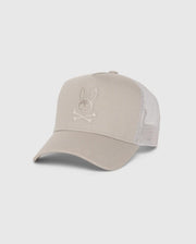 The MENS MALTON TRUCKER CAP - B6A797D200 by Psycho Bunny is a beige cap with a curved brim and mesh back for ventilation. Made from 100% cotton, it features an embroidered bunny-and-crossbones design on the front.
