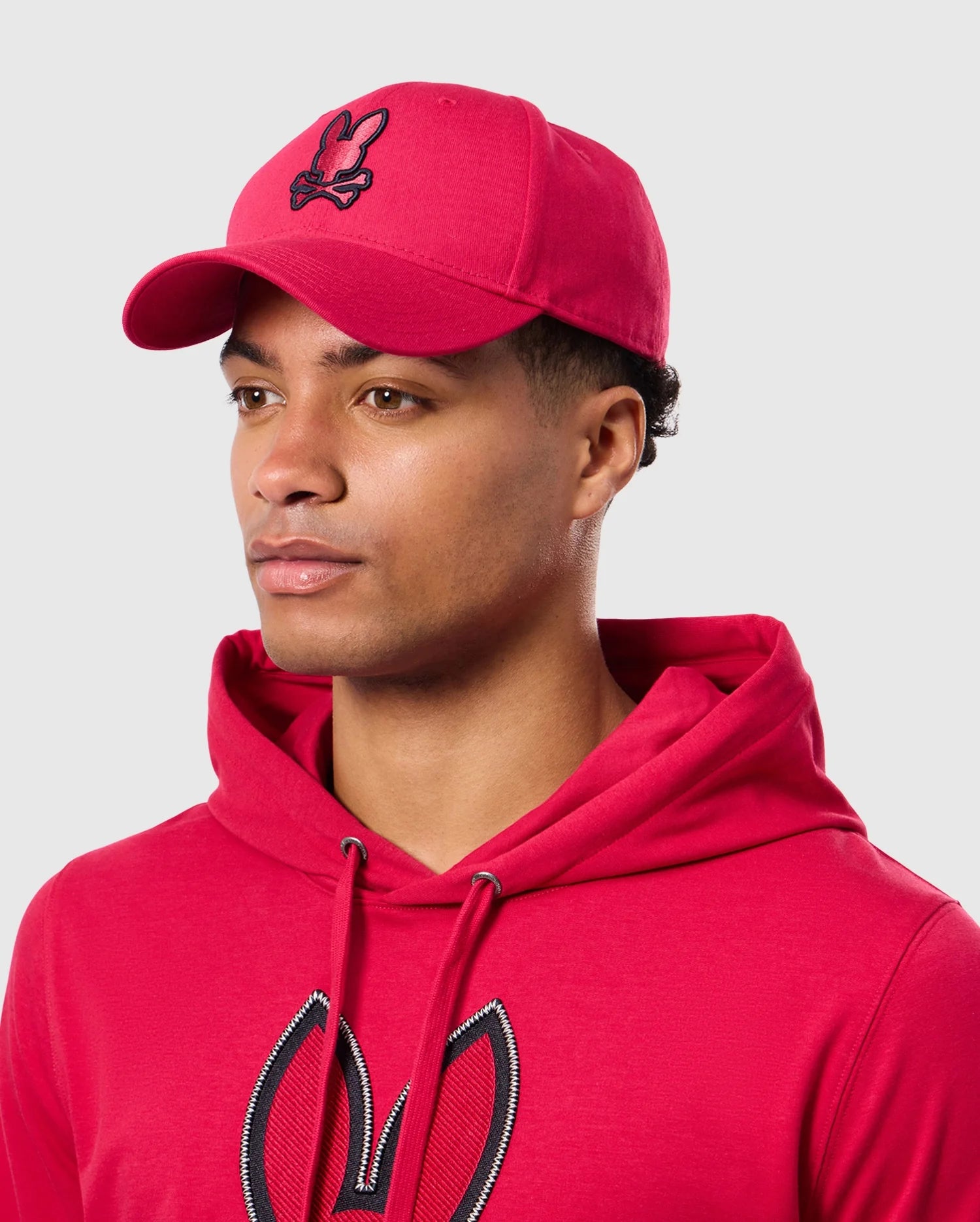 A person wearing a Psycho Bunny red MENS WALTER BASEBALL CAP - B6A800D200 and a hoodie with a stylized rabbit logo. Made from 100% cotton for durability and comfort, the outfit is set against a plain light grey background as the individual looks sideways with a neutral expression.