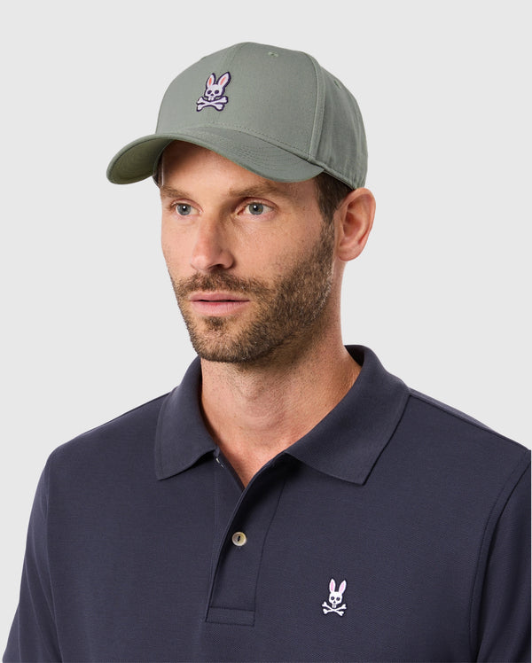 A man in a Psycho Bunny MENS CLASSIC BASEBALL CAP - B6A816B200 with a bunny and crossbones logo, paired with a navy polo shirt featuring the same emblem. With short hair and a beard, he stands against a light gray background, embodying effortless style in men's accessories.