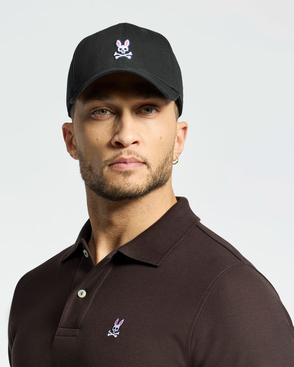 A person wears a Psycho Bunny MENS CLASSIC BASEBALL CAP - B6A816A1HT in black with an adjustable fit and bunny skull logo, paired with a brown polo. They have a beard, hoop earring, and look directly at the camera against a plain background.