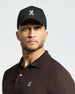 A person wears a Psycho Bunny MENS CLASSIC BASEBALL CAP - B6A816A1HT in black with an adjustable fit and bunny skull logo, paired with a brown polo. They have a beard, hoop earring, and look directly at the camera against a plain background.