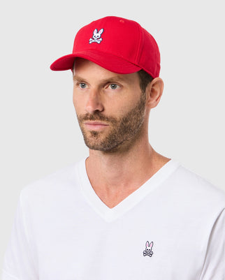 A bearded man sports a Psycho Bunny MENS CLASSIC BASEBALL CAP - B6A816A1HT in classic red, featuring an embroidered skull and bunny logo. His white V-neck T-shirt matches with the same emblem against a plain gray backdrop, highlighting his stylish look.