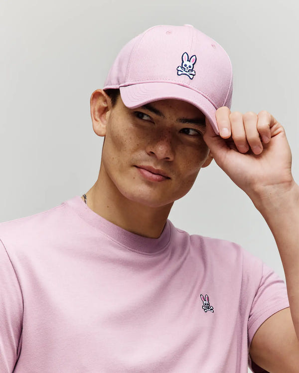 A person wears a light pink Psycho Bunny MENS CLASSIC BASEBALL CAP (B6A816A1HT) and matching T-shirt, both featuring a cartoon bunny and crossbones logo, looking to the side while holding the brim. The background is plain and light.