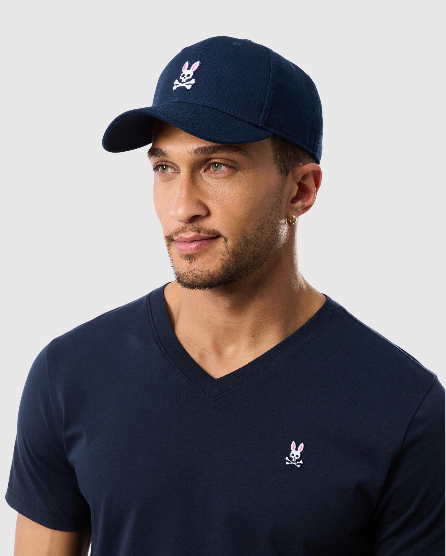 A man wears a Psycho Bunny navy baseball cap (B6A816A1HT) with an embroidered pink bunny logo and a matching V-neck T-shirt, both in soft cotton twill. He has a short beard, a hoop earring, and stands against a plain light gray background.