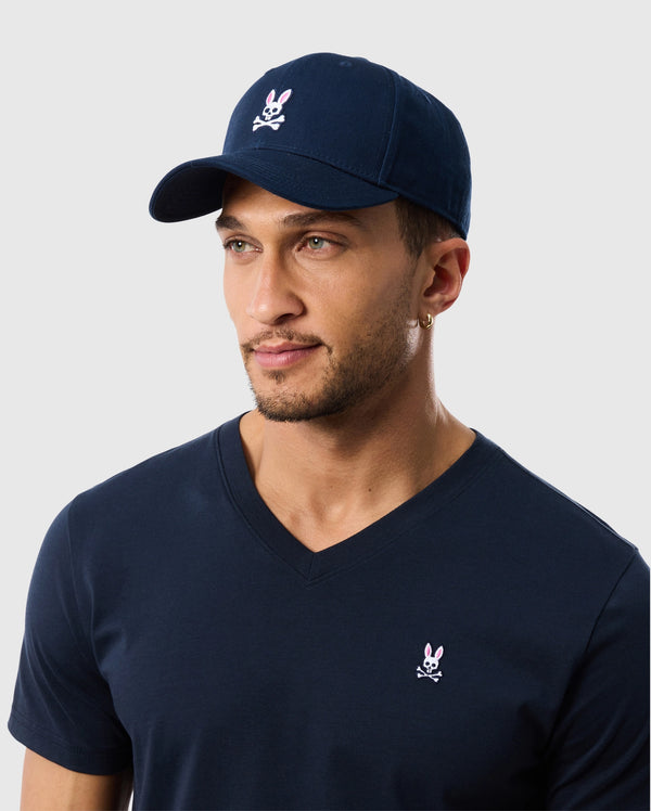 A man wears a Psycho Bunny navy baseball cap (B6A816A1HT) with an embroidered pink bunny logo and a matching V-neck T-shirt, both in soft cotton twill. He has a short beard, a hoop earring, and stands against a plain light gray background.