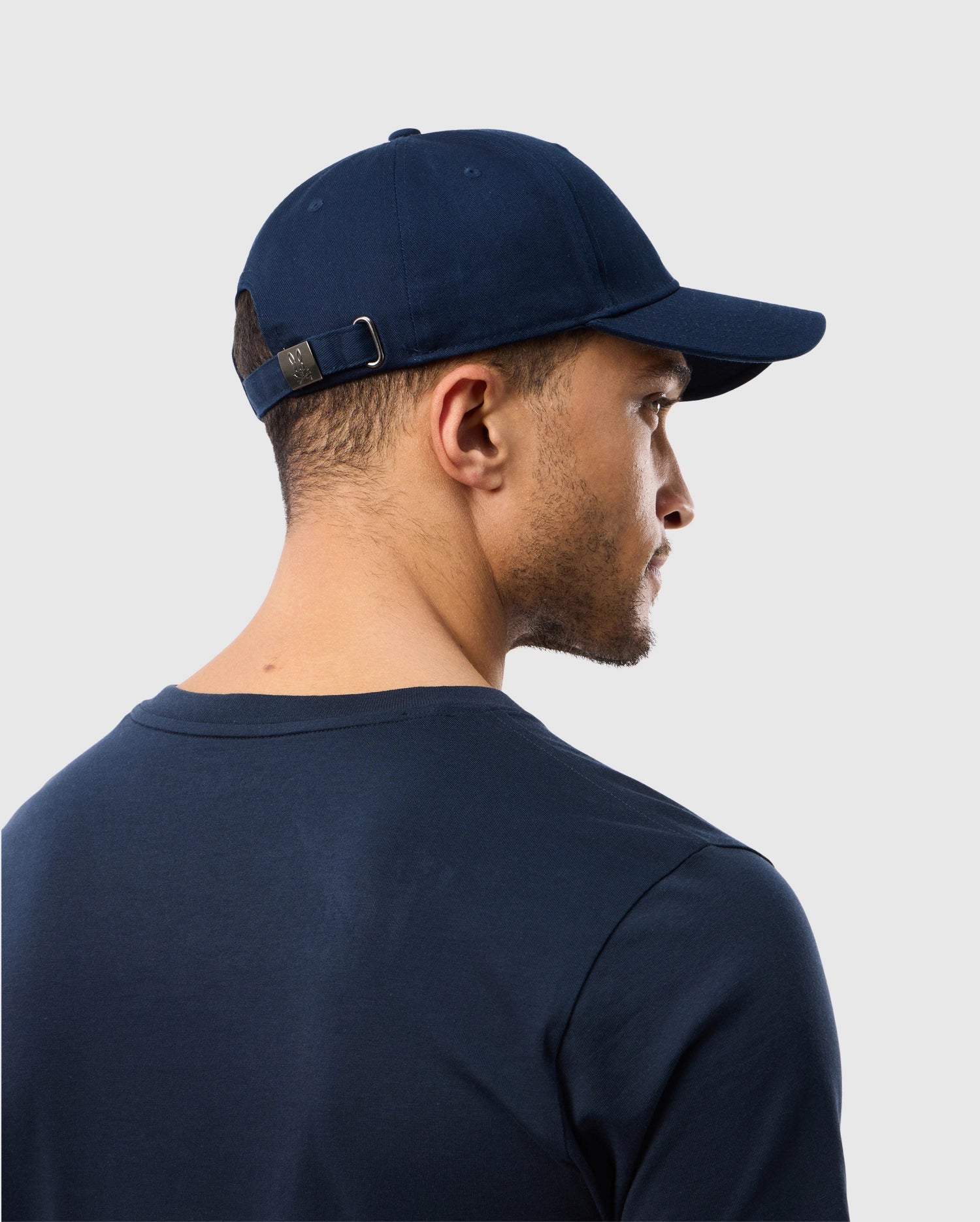 A person wearing a Psycho Bunny MENS CLASSIC BASEBALL CAP - B6A816A1HT in navy blue and a matching t-shirt is shown from behind. The cotton twill cap features an adjustable strap and subtle embroidered logo, set against a plain light-colored background.
