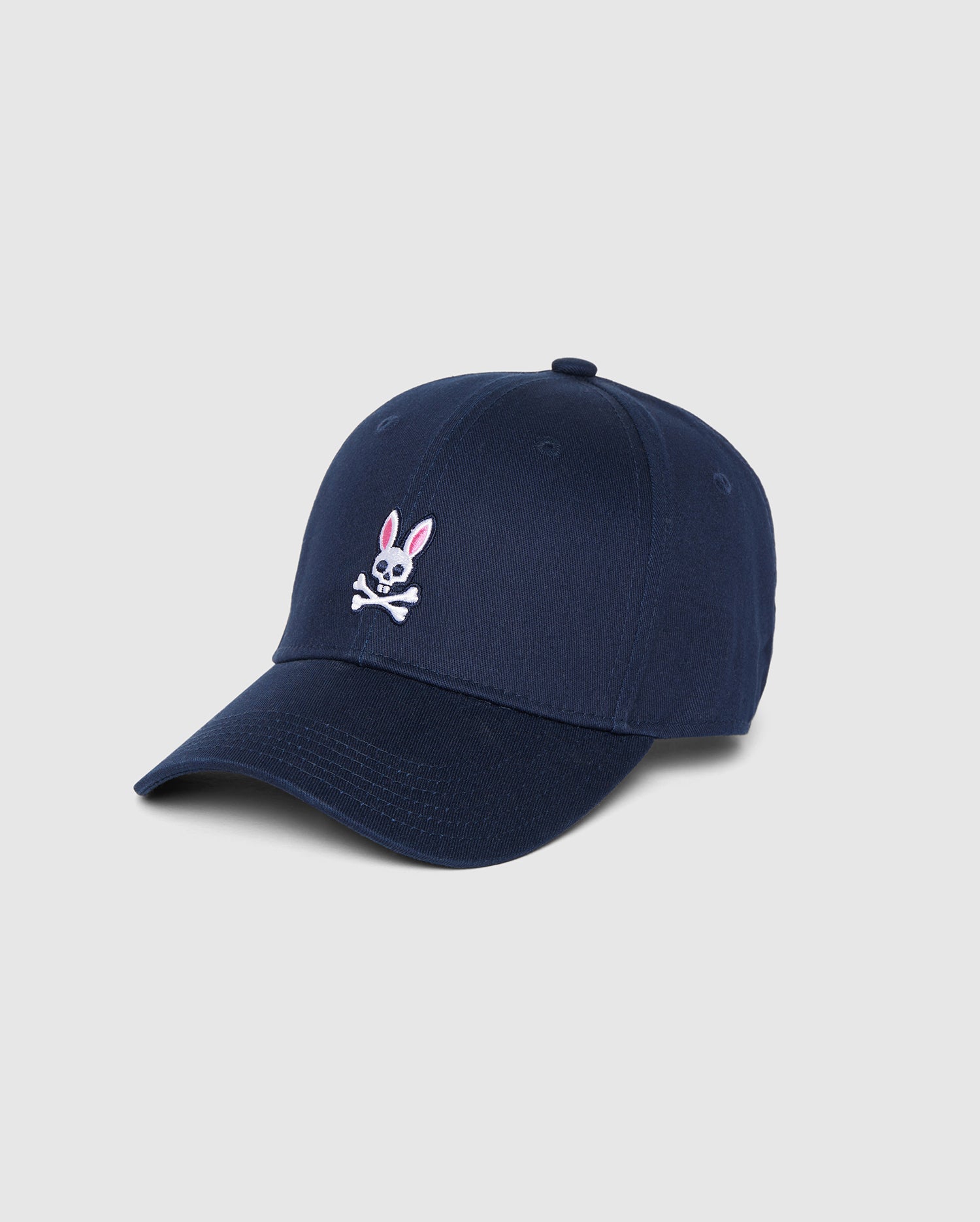 This MENS CLASSIC BASEBALL CAP - B6A816A1HT, made from cotton twill, features an embroidered design of a pink and white bunny with crossbones underneath on the front. The navy blue cap has a curved brim and an adjustable strap at the back. The background is plain white, making it a standout men's accessory by Psycho Bunny.