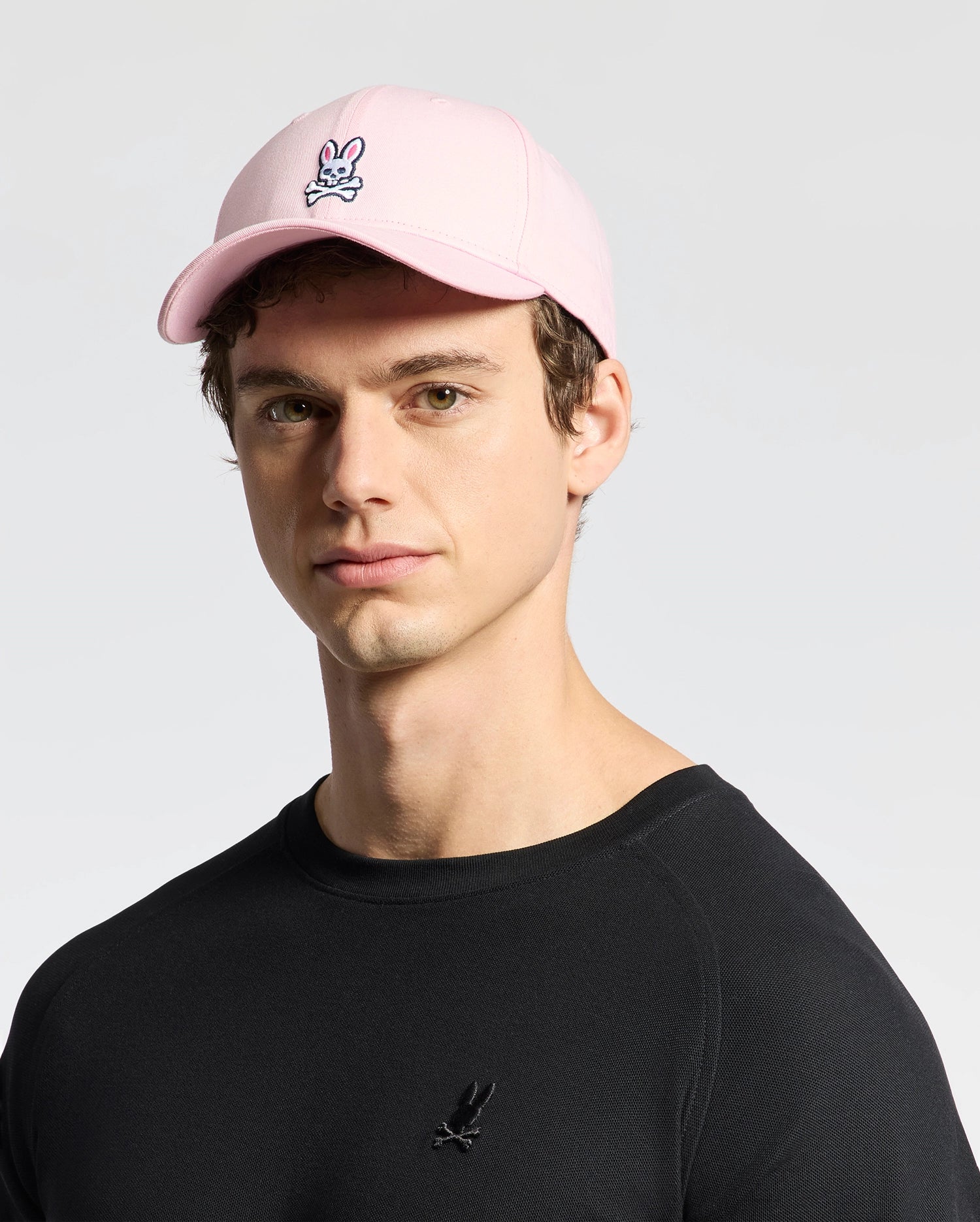 Wearing a pink cotton twill MENS CLASSIC BASEBALL CAP - B6A816A1HT by Psycho Bunny with an embroidered Bunny logo, and a black shirt with a small logo, the person maintains a neutral expression while facing the camera against a light background.