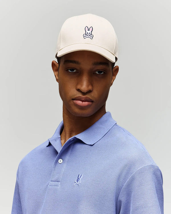 A young man in a Psycho Bunny MENS CLASSIC BASEBALL CAP - B6A816A1HT, featuring an adjustable cotton fit and embroidered Bunny and crossbones logo, wears a light purple polo shirt against a plain light gray background.