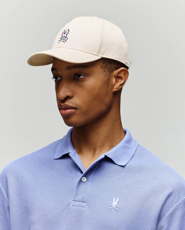 A man wearing a Psycho Bunny MENS CLASSIC BASEBALL CAP (B6A816A1HT) and a light purple polo, both with the embroidered Bunny logo, looks to the side against a plain light background.