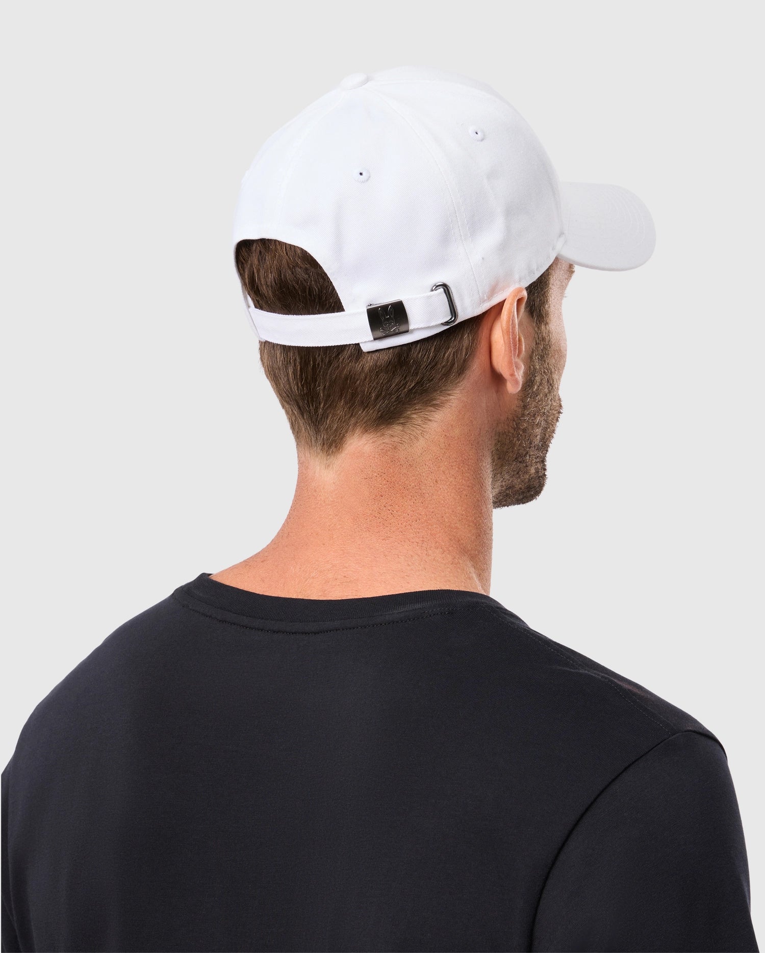 A person in a black shirt and a white Psycho Bunny MENS CLASSIC BASEBALL CAP - B6A816A1HT is shown from behind, highlighting the adjustable strap.