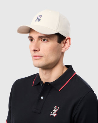 A person is stylishly dressed in a Psycho Bunny ensemble—a beige MENS CLASSIC BASEBALL CAP - B6A816B200, 100% cotton, and a black polo shirt. Both feature the rabbit graphic. The subtle gray background enhances the casually elegant look.