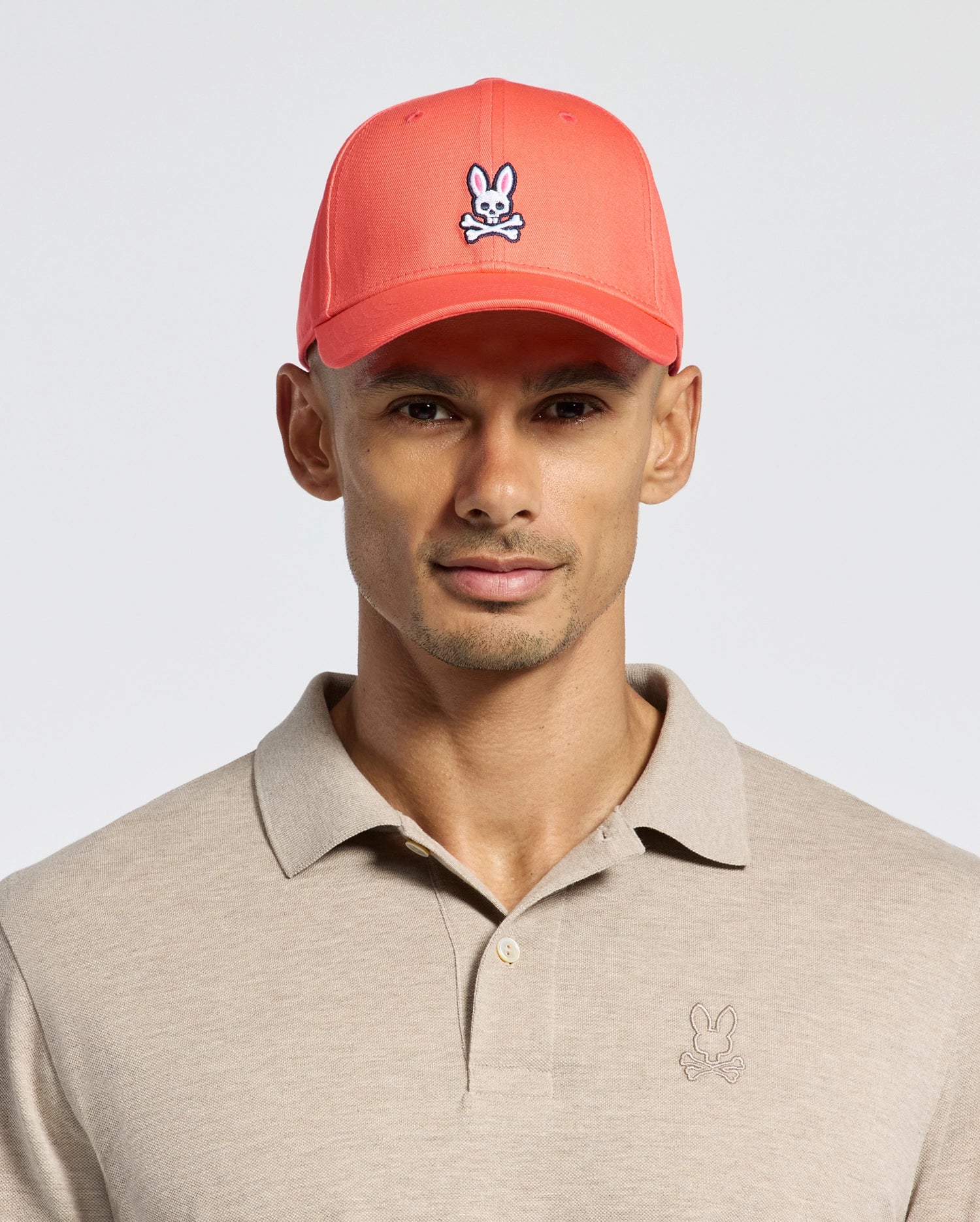 Wearing Psycho Bunny's MENS CLASSIC BASEBALL CAP, B6A816F200, in coral pink with a cartoon bunny logo in cotton twill and an adjustable fit, paired with a beige polo featuring the same logo, all set against a plain light background.