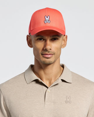 Wearing Psycho Bunny's MENS CLASSIC BASEBALL CAP, B6A816F200, in coral pink with a cartoon bunny logo in cotton twill and an adjustable fit, paired with a beige polo featuring the same logo, all set against a plain light background.