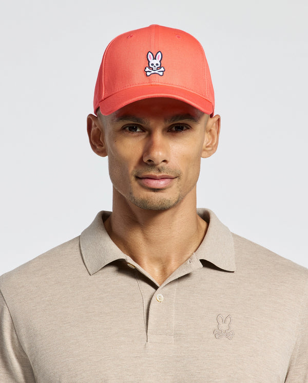 Wearing Psycho Bunny's MENS CLASSIC BASEBALL CAP, B6A816F200, in coral pink with a cartoon bunny logo in cotton twill and an adjustable fit, paired with a beige polo featuring the same logo, all set against a plain light background.