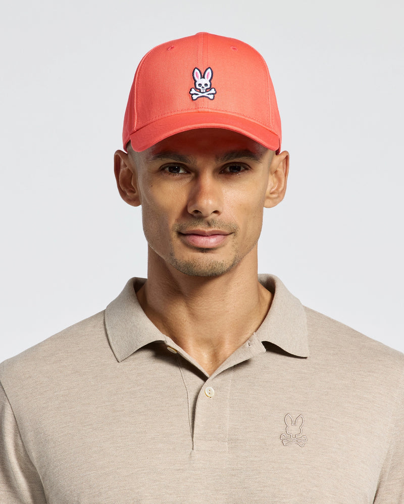 Wearing Psycho Bunny's MENS CLASSIC BASEBALL CAP, B6A816F200, in coral pink with a cartoon bunny logo in cotton twill and an adjustable fit, paired with a beige polo featuring the same logo, all set against a plain light background.