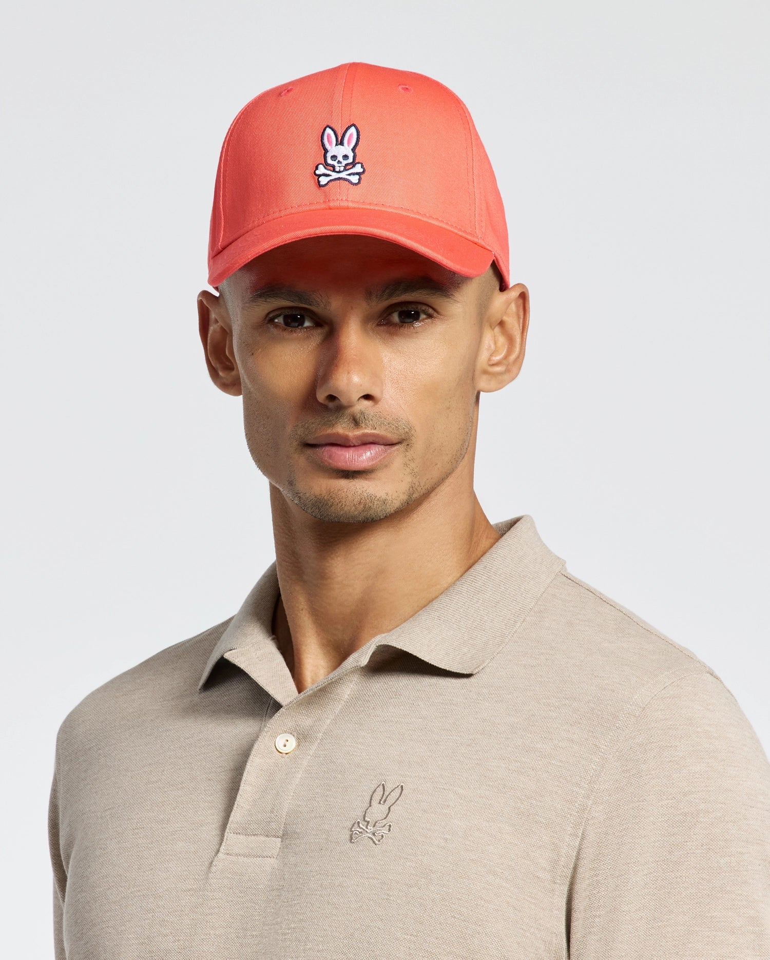 A person wears a Psycho Bunny coral baseball cap with a rabbit and crossbones logo, offering an adjustable fit. They pair it with a classic beige polo shirt featuring the same small rabbit logo. The cotton twill cap contrasts nicely against the neutral background.