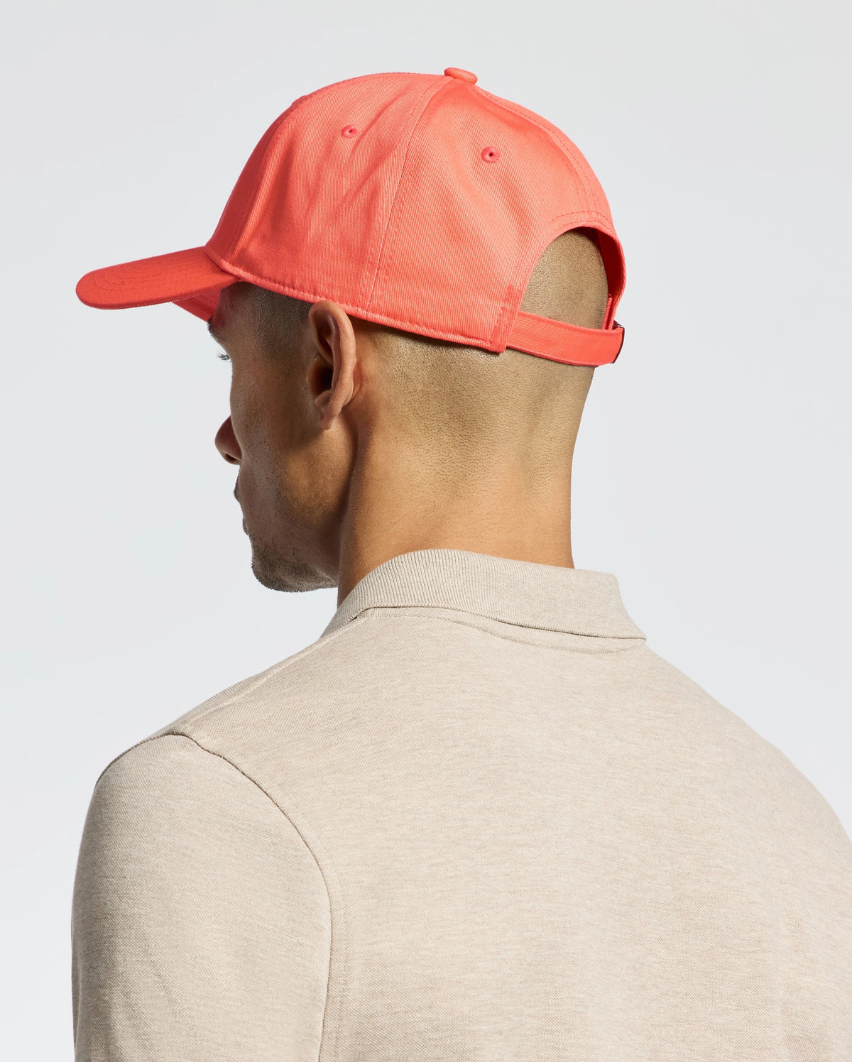 A person in a coral-colored Psycho Bunny MENS CLASSIC BASEBALL CAP - B6A816F200 is viewed from the side and back. Dressed in a beige shirt, they maintain a neutral expression and classic style against a plain, light-colored background.