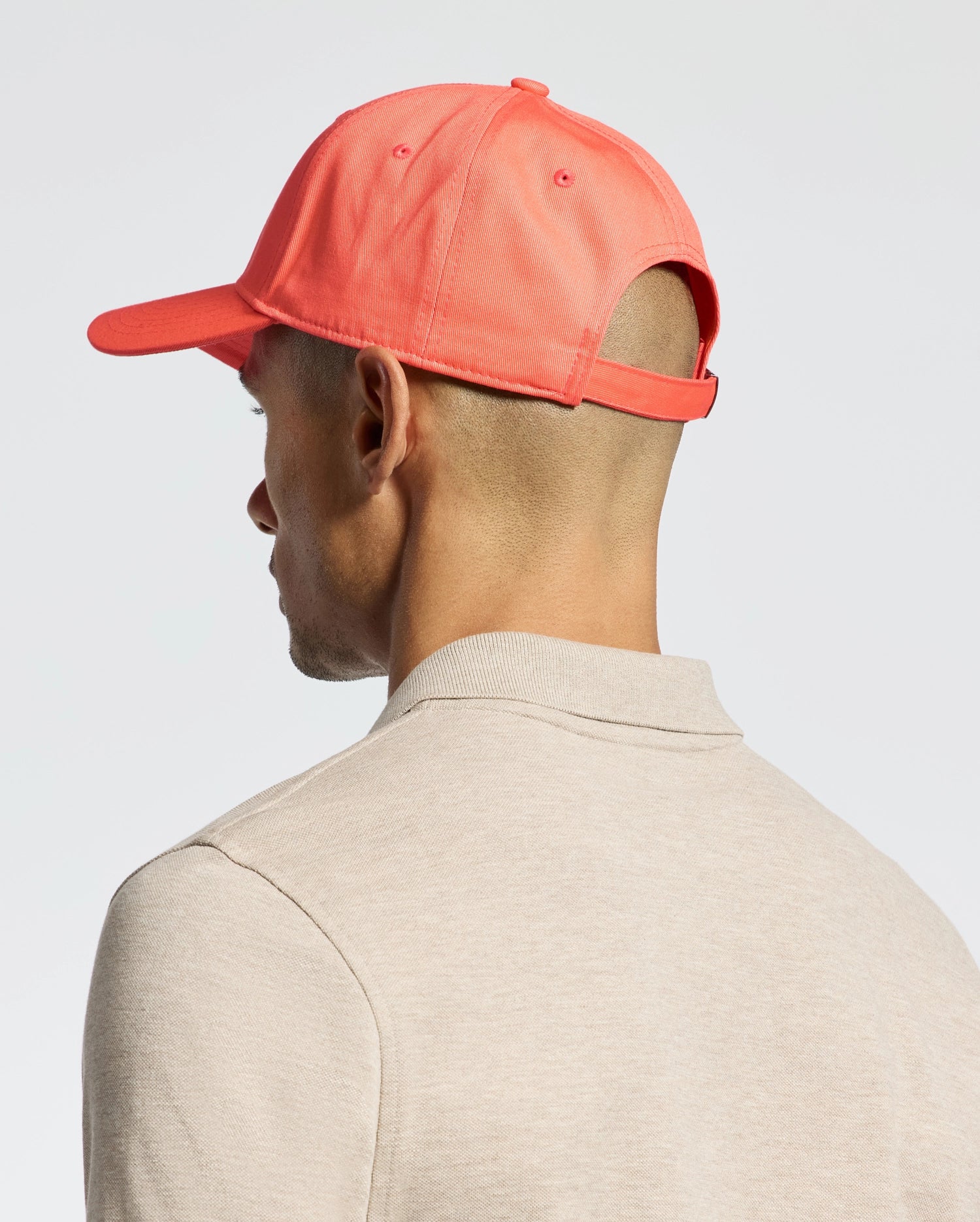 A person in a coral-colored Psycho Bunny MENS CLASSIC BASEBALL CAP - B6A816F200 is viewed from the side and back. Dressed in a beige shirt, they maintain a neutral expression and classic style against a plain, light-colored background.