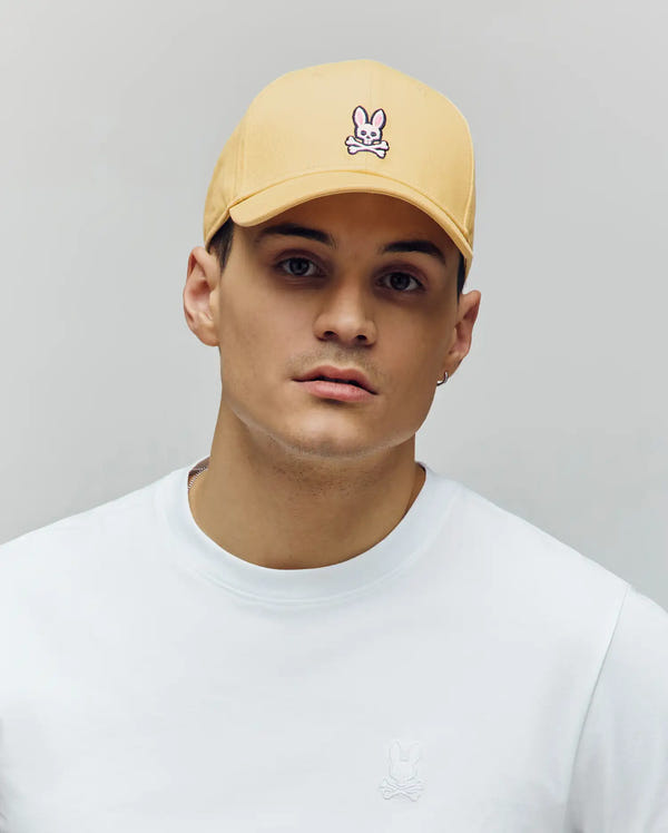 A young man in a Psycho Bunny MENS CLASSIC BASEBALL CAP (B6A816F200), pale yellow, made from recycled polyester with a bunny logo and adjustable fit, pairs it with a white crew-neck shirt, standing against a light gray background.