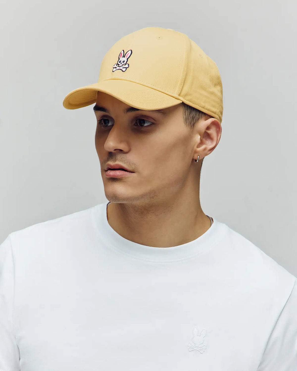 A young man in a Psycho Bunny MENS CLASSIC BASEBALL CAP - B6A816F200 (light yellow, small bunny logo), a white recycled polyester t-shirt, and a small hoop earring looks slightly to the side against a plain background.