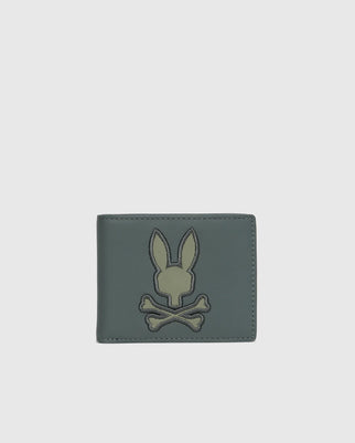 Introducing the MENS WORDMARK LEATHER WALLET - B6A868D200 from Psycho Bunny: A stylish dark green leather wallet showcasing a stitched design of a bunny head above two crossed bones. This contemporary leather accessory is displayed closed against a light gray background, ideal for the fashionable man.