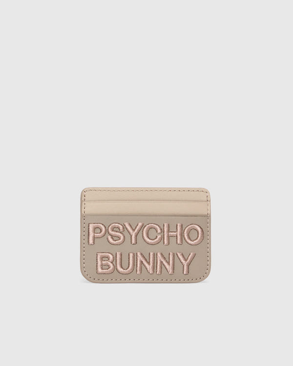 Beige leather card holder from Psycho Bunny with "PSYCHO BUNNY" in pink embroidery. The MENS WORDMARK LEATHER CARD CASE - B6A869D200 features a minimalist design on a pure white background, highlighting its premium leather craftsmanship.