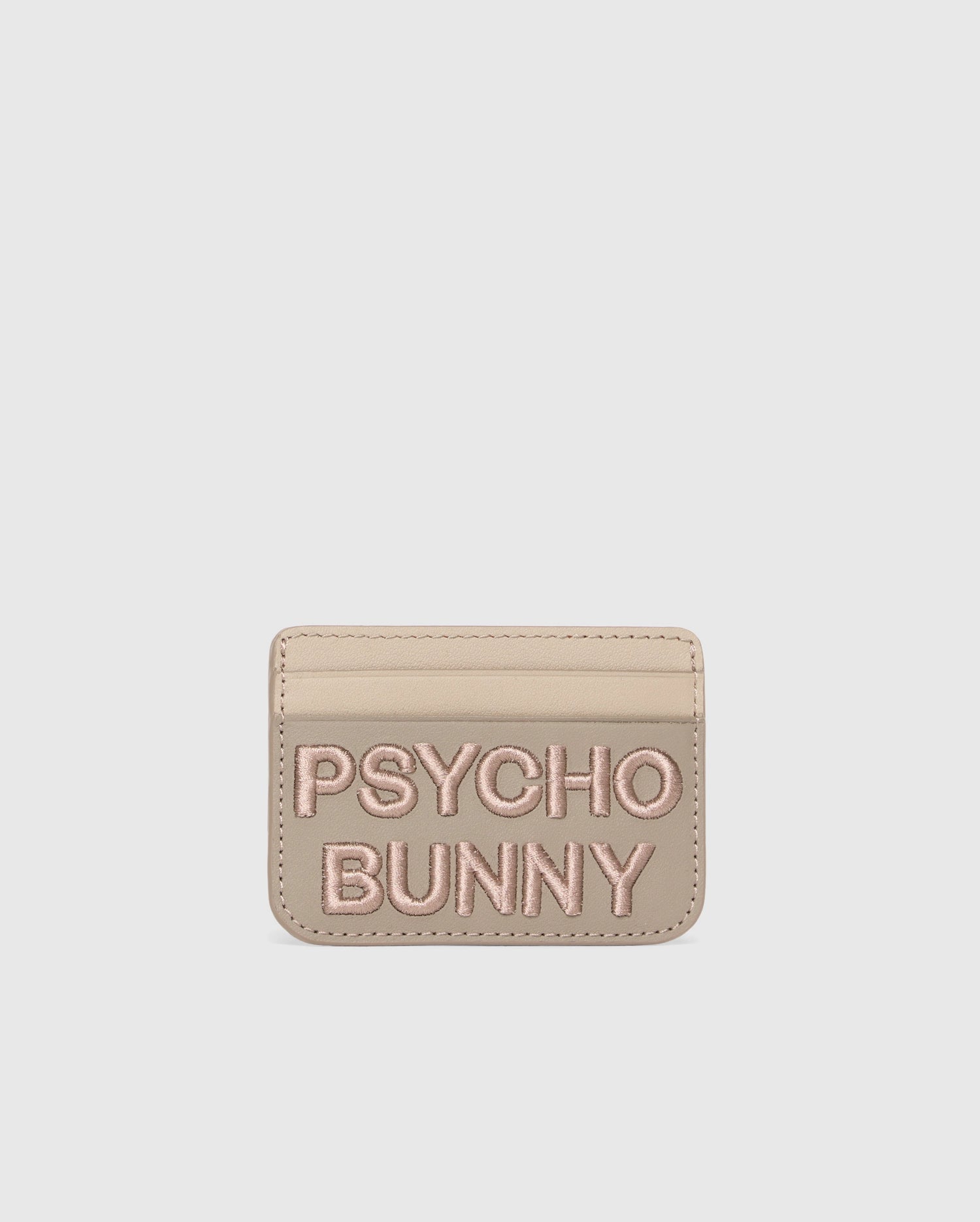 Beige leather card holder from Psycho Bunny with "PSYCHO BUNNY" in pink embroidery. The MENS WORDMARK LEATHER CARD CASE - B6A869D200 features a minimalist design on a pure white background, highlighting its premium leather craftsmanship.