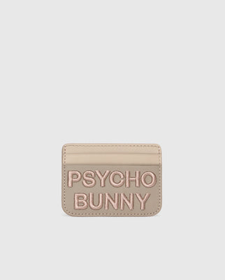 Beige leather card holder from Psycho Bunny with "PSYCHO BUNNY" in pink embroidery. The MENS WORDMARK LEATHER CARD CASE - B6A869D200 features a minimalist design on a pure white background, highlighting its premium leather craftsmanship.