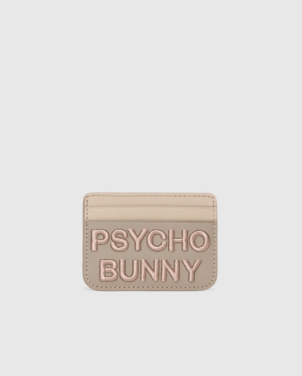 Beige leather card holder from Psycho Bunny with "PSYCHO BUNNY" in pink embroidery. The MENS WORDMARK LEATHER CARD CASE - B6A869D200 features a minimalist design on a pure white background, highlighting its premium leather craftsmanship.