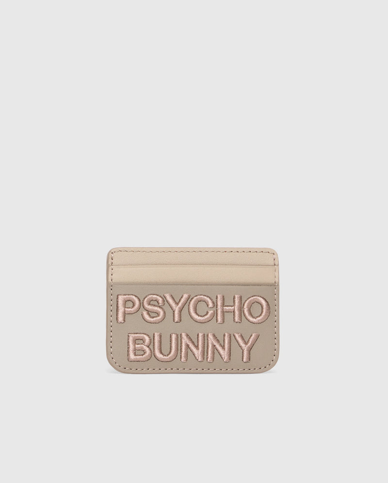 Beige leather card holder from Psycho Bunny with "PSYCHO BUNNY" in pink embroidery. The MENS WORDMARK LEATHER CARD CASE - B6A869D200 features a minimalist design on a pure white background, highlighting its premium leather craftsmanship.
