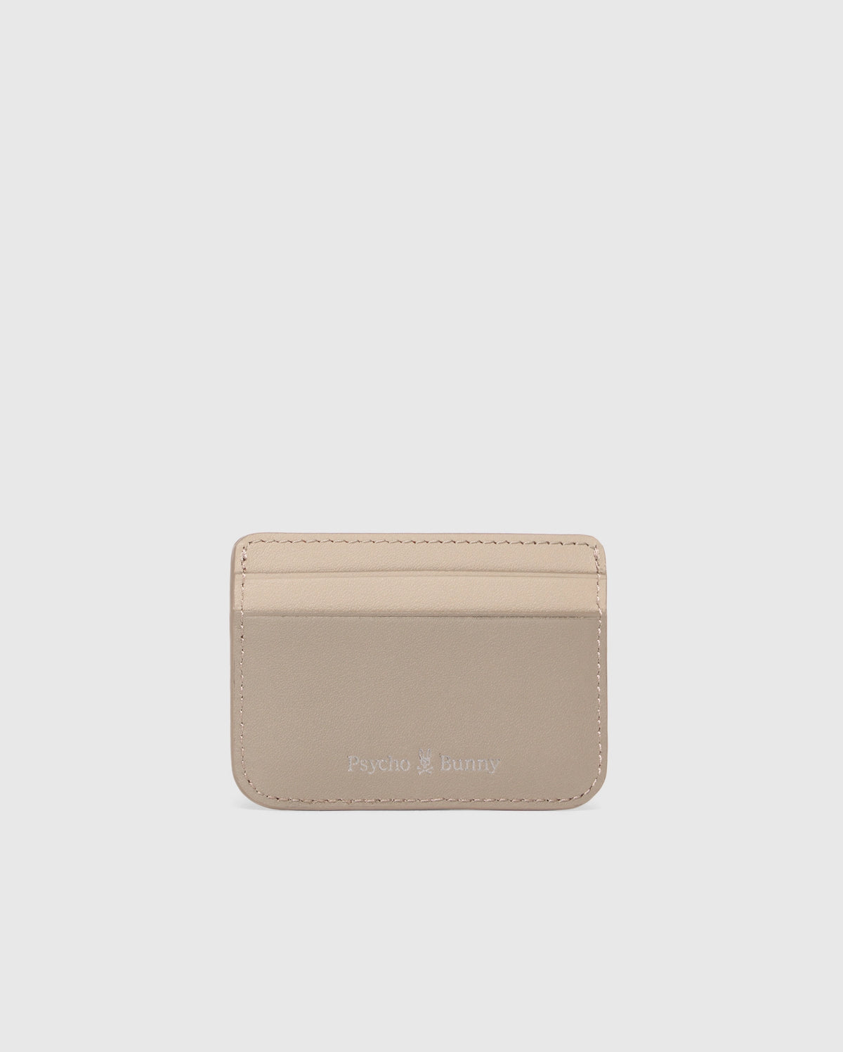 The MENS LEATHER CARD CASE - B6A869D200 by Psycho Bunny is a premium beige leather cardholder with three front slots and a silver "Psycho Bunny" embossing near the bottom center, minimal edge stitching on a plain light gray background.